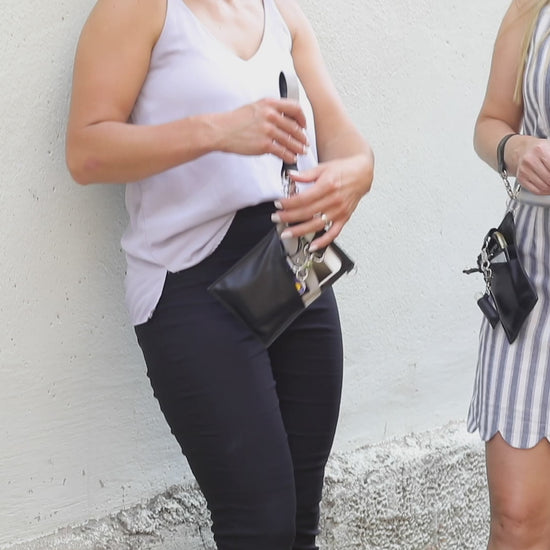 A person with a black vegan leather phone pouch with a black vegan leather magnetic wristlet key chain and a black removable three pocket wallet and black lip balm holder set. Another Person  with a black and gray vegan leather bracelet, keychain, three pocket wallet, and a black lip balm holder
